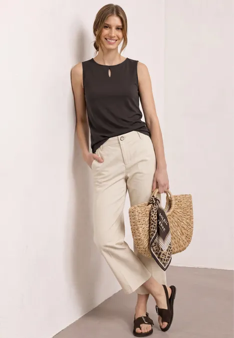 Frau in schwarzem Top und beiger Hose, mit schwarzen Sandalen und Strohtasche mit Tuch.