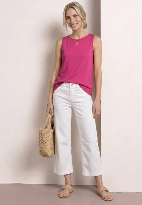 Frau trägt pinkes ärmelloses Top, weiße Culotte, Basttasche und Sandalen vor hellem Hintergrund.