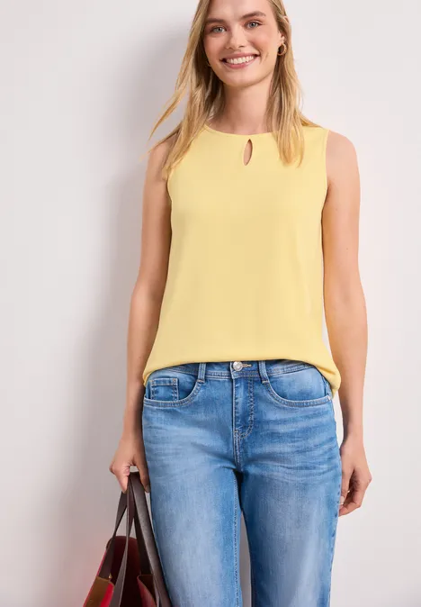 Frau in gelbem ärmellosem Top und blauen Jeans hält eine Handtasche.