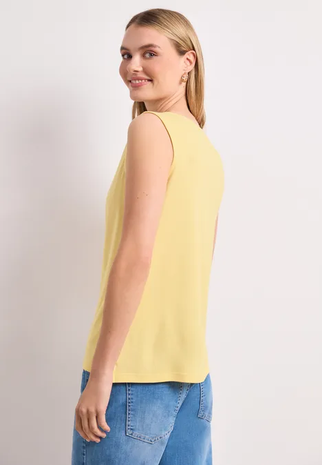 Eine Frau trägt ein gelbes ärmelloses Top und blaue Jeans, seitlich stehend vor weißem Hintergrund.