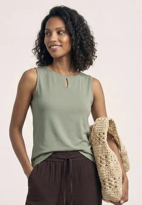Frau mit lockigem Haar trägt grünes ärmelloses Top, braune Hose und eine gehäkelte Tasche.