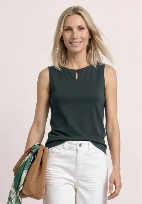 Frau in dunkelgrünem ärmellosem Top, weißer Hose und brauner Schultertasche.