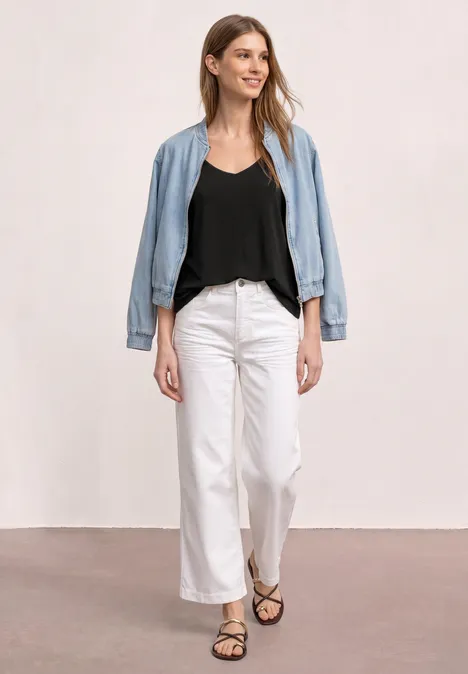 Eine Frau in schwarzem Top, weißer Hose, offen getragener Jeansjacke und Sandalen vor heller Wand.