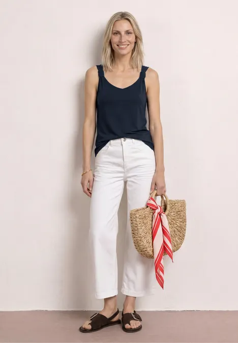 Top met V-hals en knoopdetail deep blue Frau in ärmellosem Top, weißen Hosen, schwarzen Sandalen, mit Strohtasche und rotem Tuch steht vor hellem Hintergrund.