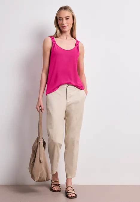 Frau trägt pinkes Top, beige Hose, Sandalen, hält große beige Tasche, vor weißem Hintergrund stehend.