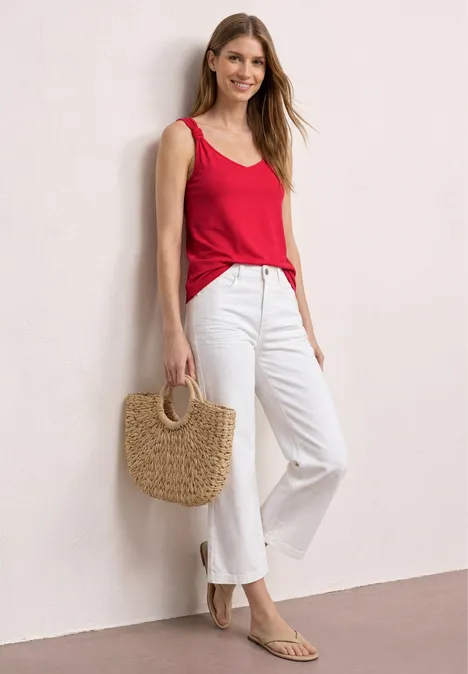 Top met V-hals en knoopdetail salsa red Frau lehnt lässig an Wand, trägt rotes Top, weiße Hose, Strohtasche und flache Sandalen.
