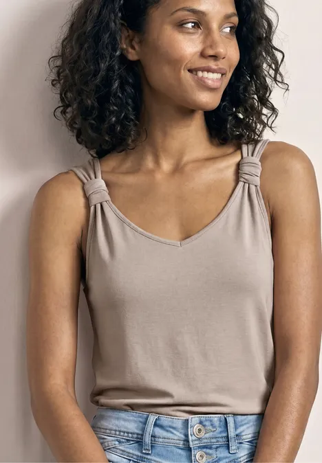 Beige ärmelloses Top mit eingedrehten Trägern, blaue Jeans, Person vor neutralem Hintergrund.