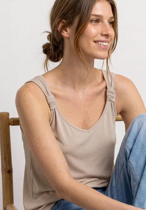 Frau sitzt lässig auf einem Stuhl, trägt ein beiges Top mit Knoten an den Trägern und blaue Jeans.