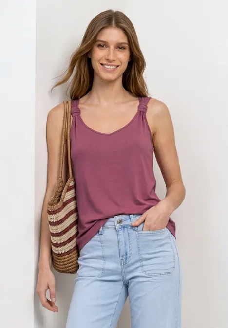 Vrouw in mouwloos bordeaux topje en spijkerbroek, met gevlochten stro-tas over de schouder.
