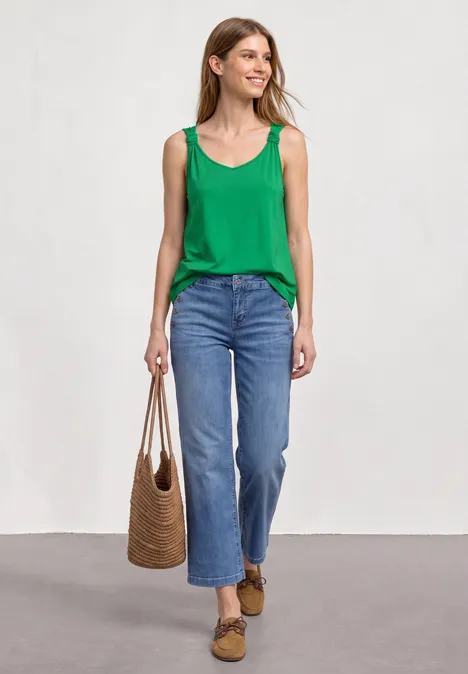 Frau in grünem Top, blauen Jeans, mit Korbtasche und braunen Slippern vor hellem Hintergrund.