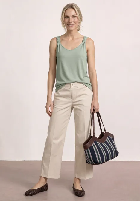 Een vrouw in een groen hemdje en beige broek, met een gestreepte tas en bruine schoenen.