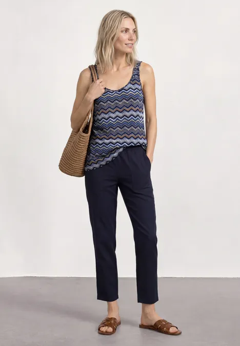Frau mit Strohtasche, gemustertem Top, dunkelblauen Hosen und braunen Sandalen.