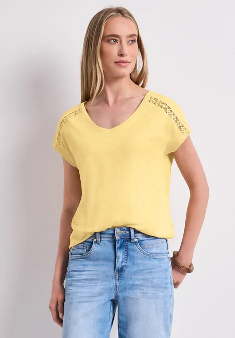 Frau in gelbem T-Shirt mit V-Ausschnitt und blauen Jeans steht vor neutralem Hintergrund.