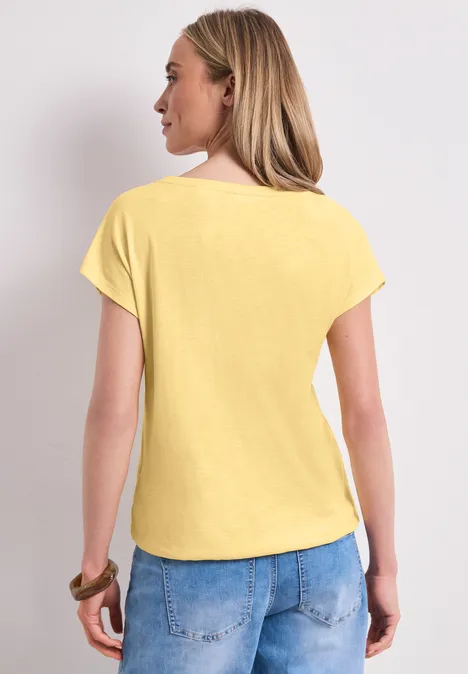 Rückansicht einer Frau in hellem Gelb-Shirt und blauer Jeans vor neutralem Hintergrund.