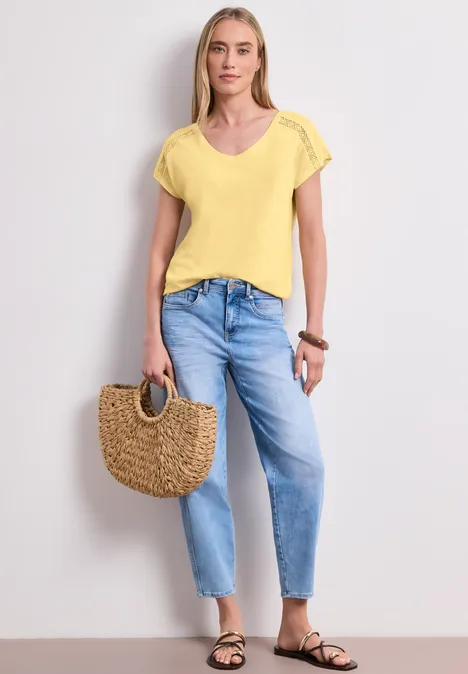 Modell in gelbem T-Shirt, weiten blauen Jeans, schwarzen Sandalen und mit runder Strohtasche vor hellem Hintergrund.