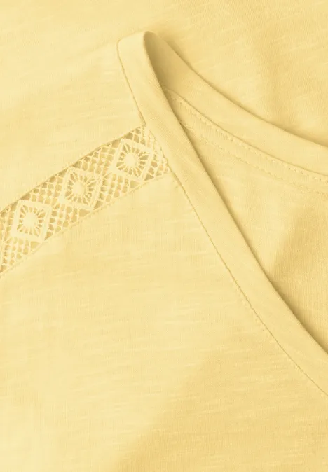 Nahaufnahme eines hellgelben T-Shirts mit V-Ausschnitt und feiner Spitzenborte an der Schulter.