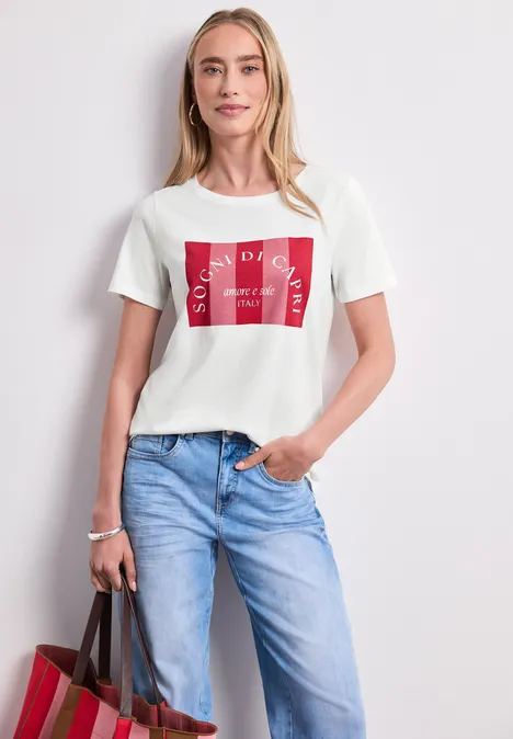 Eine Frau in blauer Jeans und weißem T-Shirt mit rotem Print, die eine Tasche trägt.