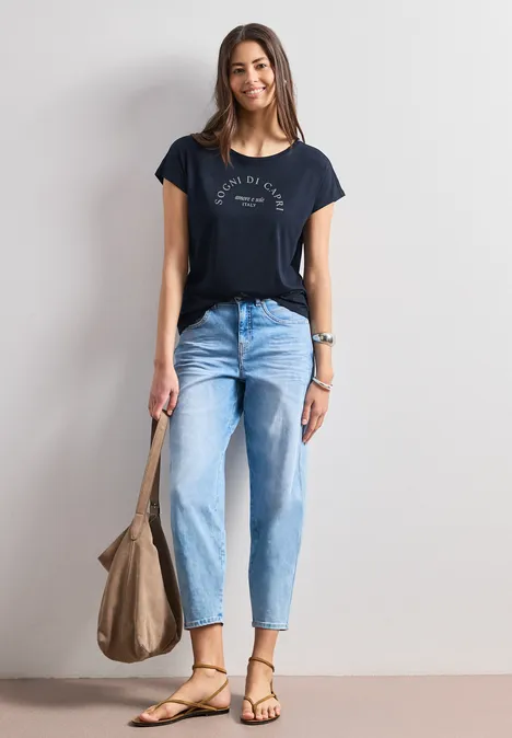 Dropped Shoulder Shirt mit Rundhals deep blue Frau vor weißer Wand in navy T-Shirt, hellen Jeans, Sandalen und mit großer Beuteltasche.