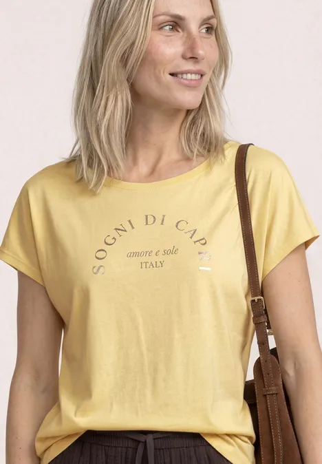 Shirt met ronde hals en losse schouders soft yellow Blonde vrouw met geel T-shirt en opschrift, glimlacht en draagt een schoudertas.