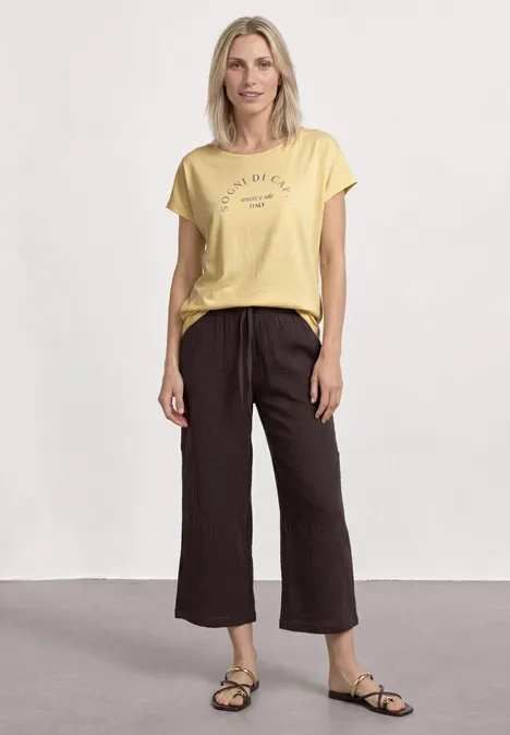 Shirt met ronde hals en losse schouders soft yellow Vrouw met geel t-shirt, wijde donkere culotte-broek en sandalen tegen lichte achtergrond.