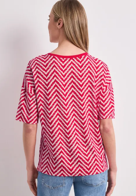 Frau von hinten in rotem-weißem Zickzack-Shirt mit halblangen Ärmeln und blauen Jeans.