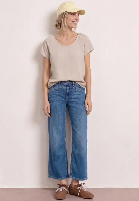 Une personne est debout devant un fond blanc, portant un t-shirt beige, un jean bleu droit et des chaussures marron.