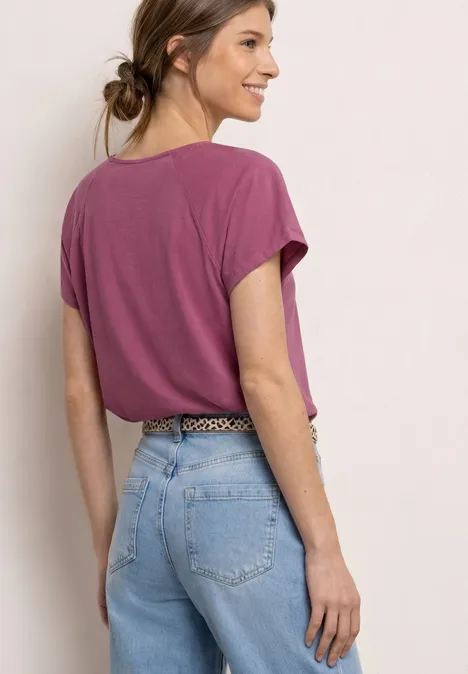 Vrouw van achteren in paars T-shirt en lichtblauwe spijkerbroek, haar nonchalant in een knot.