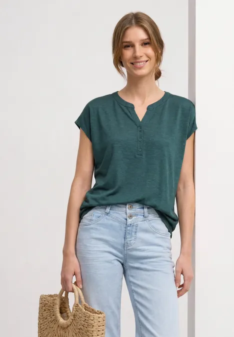 Femme portant un haut vert, un jean clair et un sac en paille sur fond neutre.