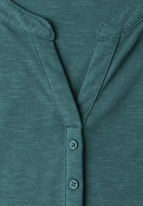 Gros plan d’un t-shirt col tunisien bleu-vert à manches longues avec patte de boutonnage et coutures décoratives.
