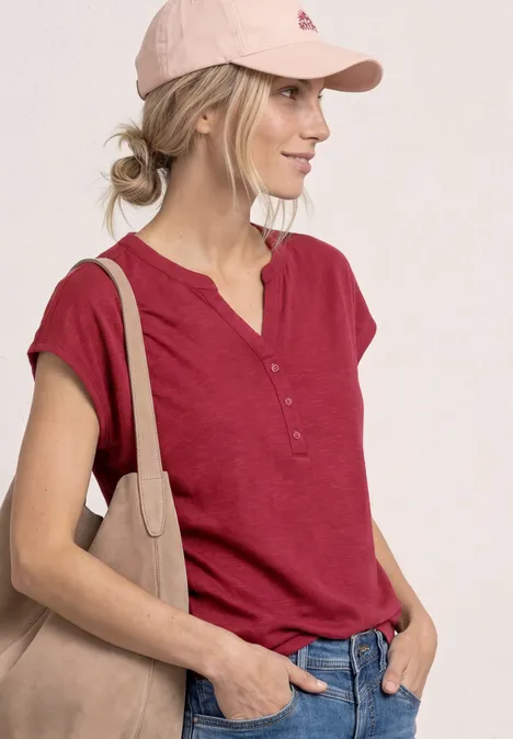Vrouw met rood kort shirt, roze pet en beige schoudertas voor een lichte achtergrond.