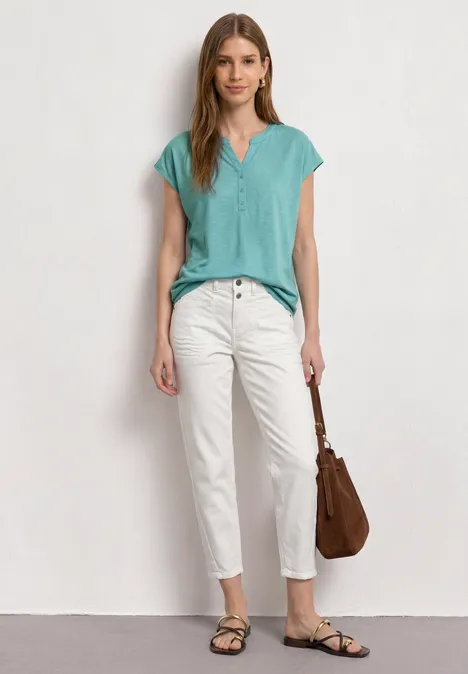 Femme debout devant un mur blanc, top turquoise, pantalon blanc, sandales noires et sac marron.