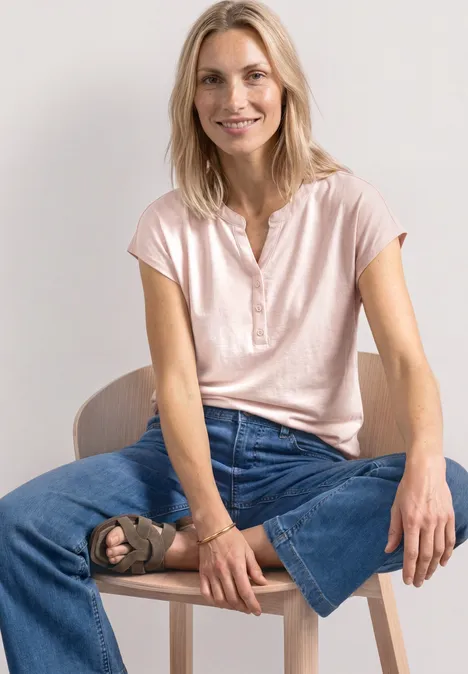 Vrouw zit op stoel met roze shirt en wijde jeans, één been opgetrokken.