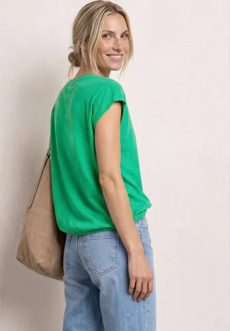 Vrouw in profiel met groen shirt, blauwe spijkerbroek en beige schoudertas voor neutrale achtergrond.