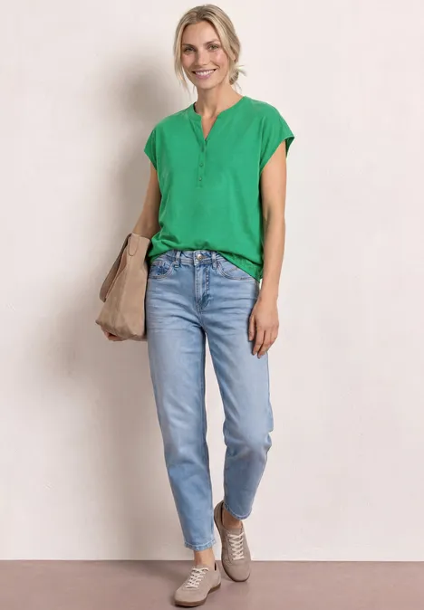 Vrouw in groen shirt en lichtblauwe jeans, met beige tas en schoenen voor lichte achtergrond.