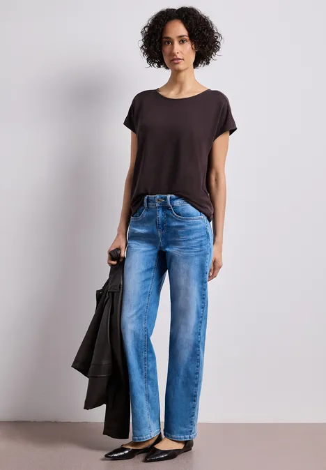 Eine Person steht mit Jeans, dunklem T-Shirt und hält eine schwarze Jacke in der Hand.