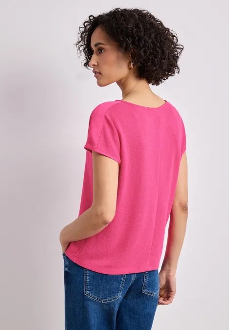 Rückansicht einer Person mit lockigen Haaren, die ein pinkes T-Shirt und Jeans vor hellem Hintergrund trägt.