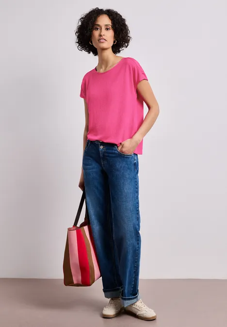 Eine Person trägt ein pinkes T-Shirt, weite blaue Jeans und hält eine gestreifte Tragetasche.