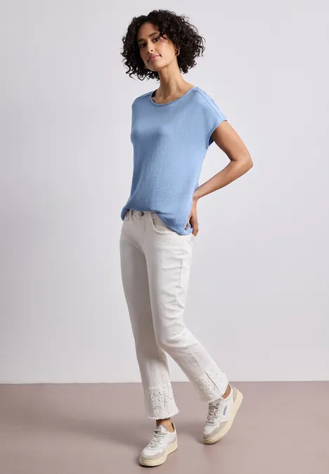 Shirt met boothals en schouderdetails drift blue Vrouw voor een neutrale achtergrond, met lichtblauw shirt, witte jeans en witte sneakers.
