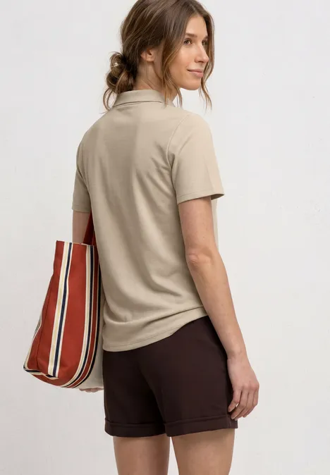 Kurzarm Poloshirt in Rippstruktur cotton beige Eine Frau steht im Profil, trägt ein beiges Poloshirt, braune Shorts und eine rot-weiß gestreifte Tasche.