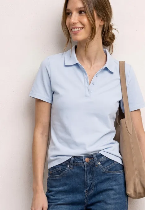 Vrouw in lichtblauw poloshirt en jeans, glimlachend met een beige schoudertas.