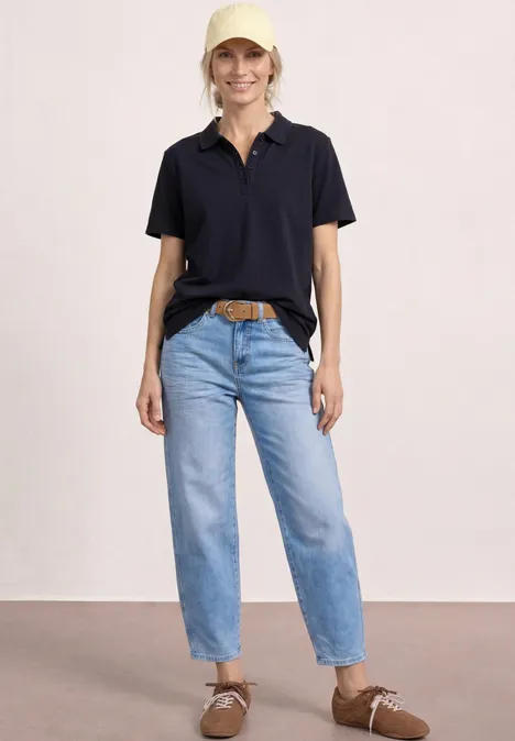 Vrouw draagt zwart poloshirt, lichtblauwe jeans met hoge taille, bruine riem en lichtbruine suède schoenen.