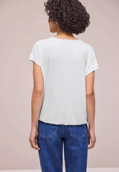 Shirt met ronde hals en decoratieve tape off white Vrouw met krullend haar van achteren, wit T-shirt en blauwe spijkerbroek, neutrale achtergrond.