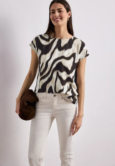 Vrouw in zebraprint top en witte jeans met een bruine hoed voor een neutrale achtergrond.