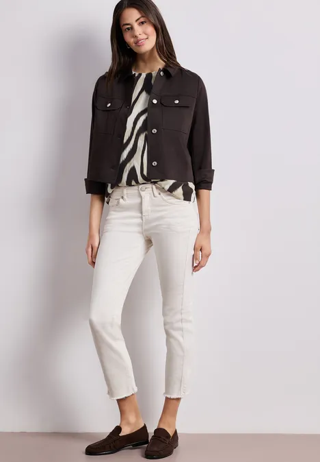 Vrouw in witte jeans, zwarte korte jas, zebra-print blouse en bruine loafers voor een lichte achtergrond.