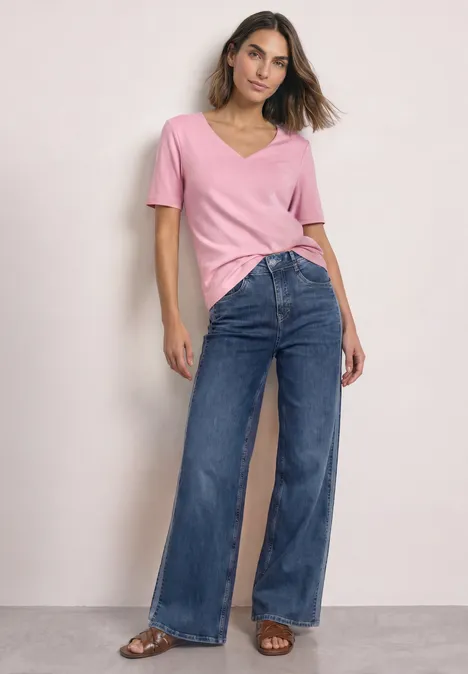 Frau in rosa V-Shirt und weiten Jeans steht vor neutralem Hintergrund.