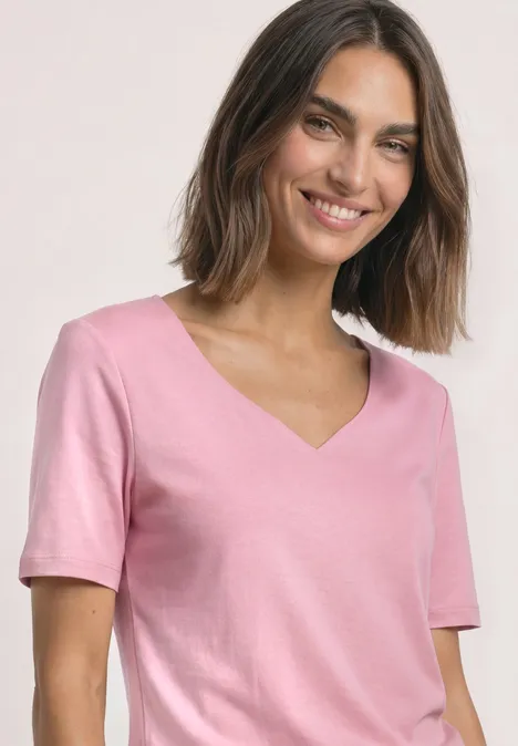 Eine lächelnde Frau trägt ein rosa T-Shirt mit V-Ausschnitt vor hellem Hintergrund.