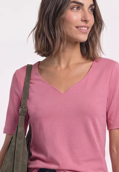 Frau in rosa V-Ausschnitt-Shirt trägt grüne Schultertasche und lächelt seitlich.
