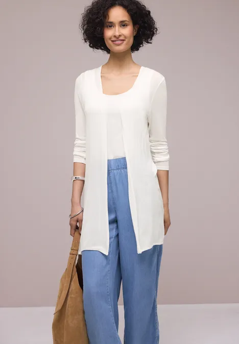 Een persoon draagt een witte open cardigan met driekwart mouwen, een blauwe wijdvallende broek en een beige tas.