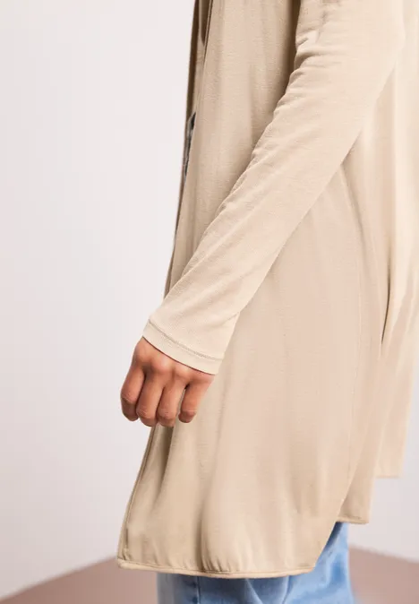 Zijaanzicht van een persoon in een lange beige cardigan en een blauwe spijkerbroek voor een lichte achtergrond.