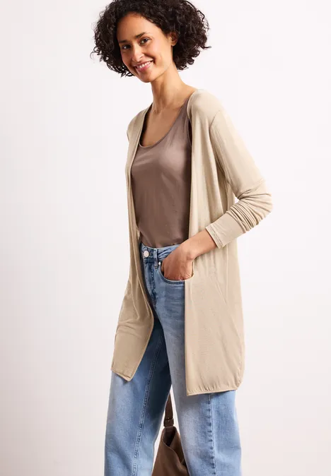 Vrouw draagt beige lange vest over bruin shirt en losse blauwe jeans, nonchalant poserend.